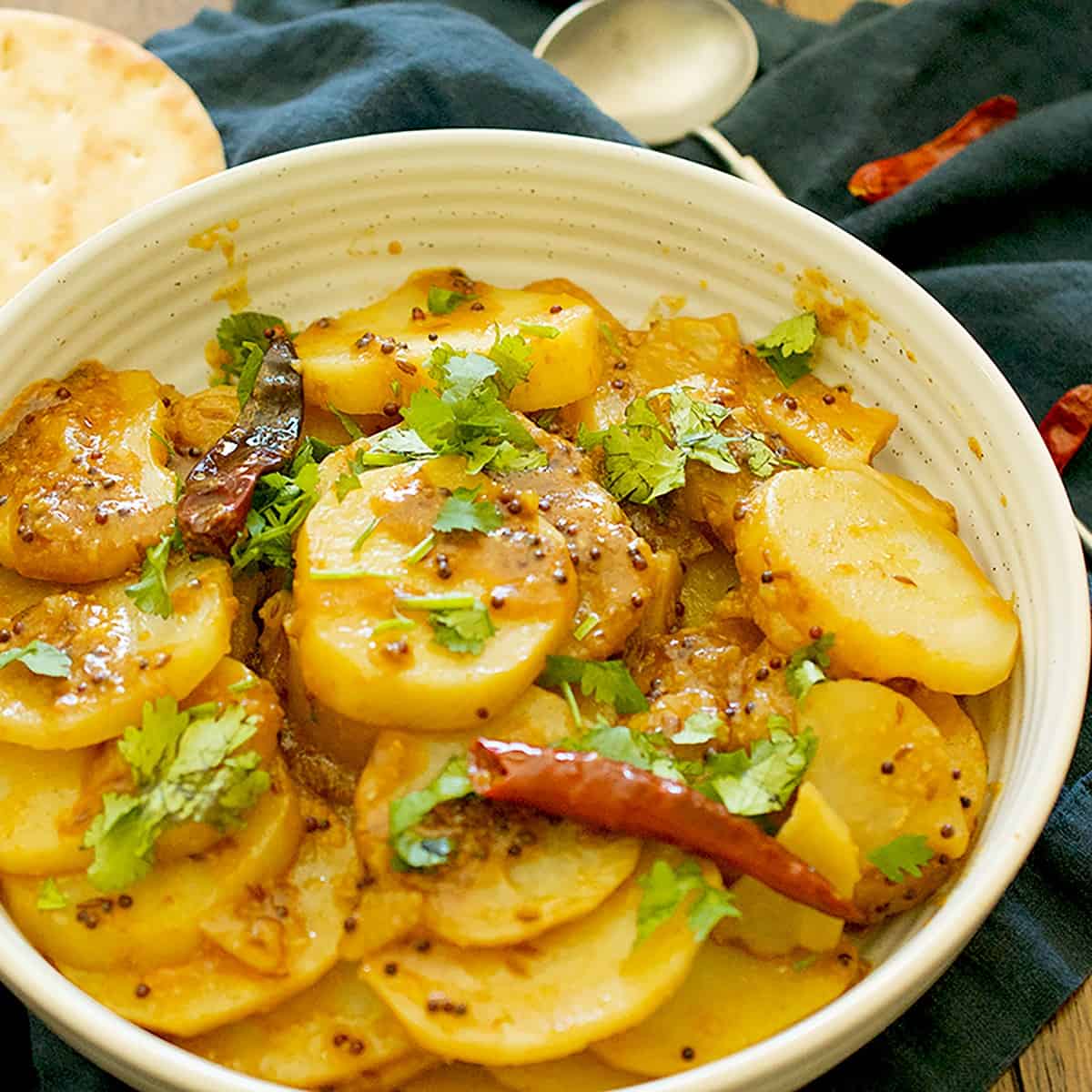 Cooked aloo bhujia, garnished with some cilantro, is plated in a white bowl and is served with some naan.