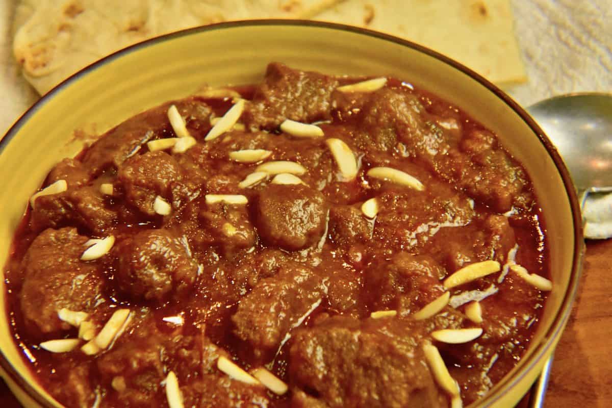 yellow bowl with gosht  & aloo bukhara topped with almonds and naan in the background.