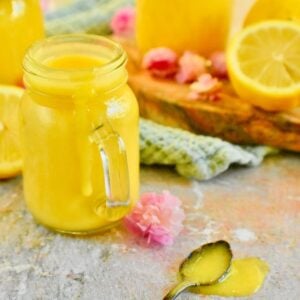 A spoon filled with some lemon curd from an easy lemon curd recipe is on a kitchen counter with some jars filled with the lemon curd, a light blue napkin, fresh lemons and pink flowers.