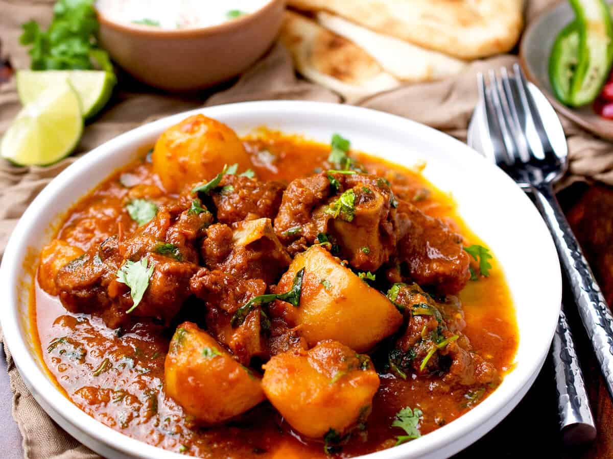 Aloo gosht recipe with naan, raita and lemons on the side.