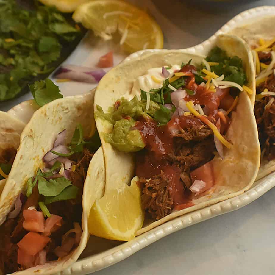 soft tacos filled with instant pot mexican shredded beef and topped with guacamole, sour cream, salsa, shredded cheese and lemons. 