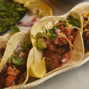 soft tacos filled with instant pot mexican shredded beef and topped with guacamole, sour cream, salsa, shredded cheese and lemons.