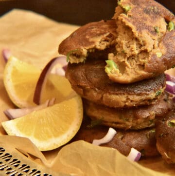 Shami kabab stacked on a silver tray lined with brown paper accompanied by lemon wedges.