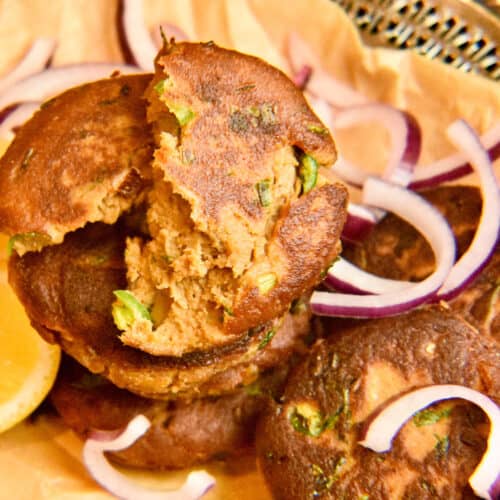 Shami kabab stacked on a silver tray lined with brown paper accompanied by lemon wedges.