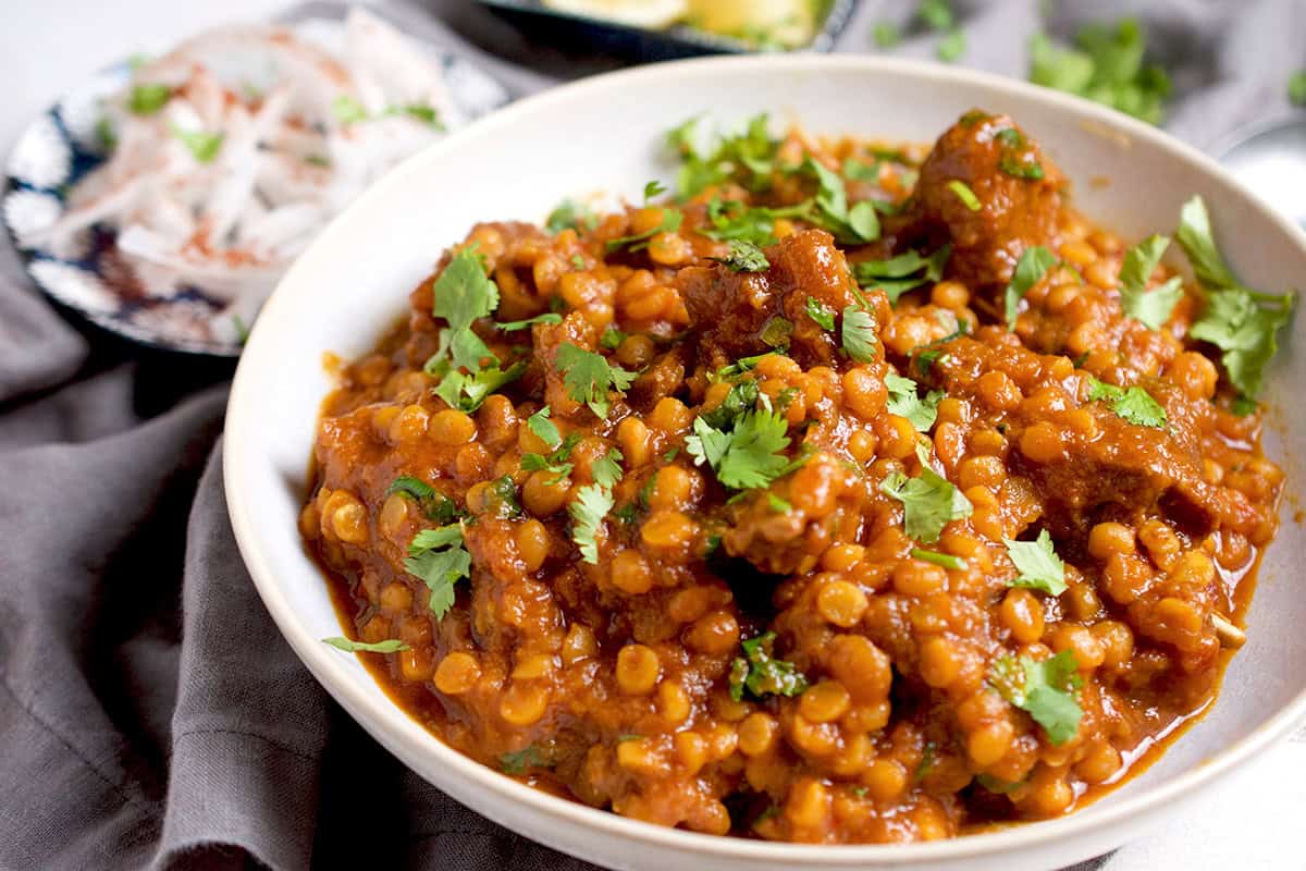 A white bowl, filled with cooked dal gosht that's garnished with 
cilantro is resting on a grey napkin with some condiments in the background.