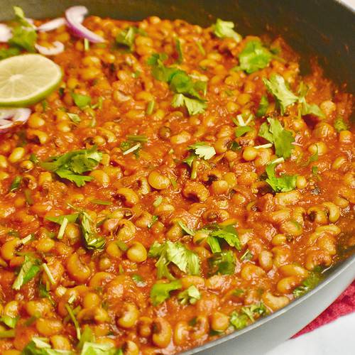 Black pan with cooked black eyed peas (Punjabi lobia) recipe, garnished with red onions, sliced lemons and cilantro.