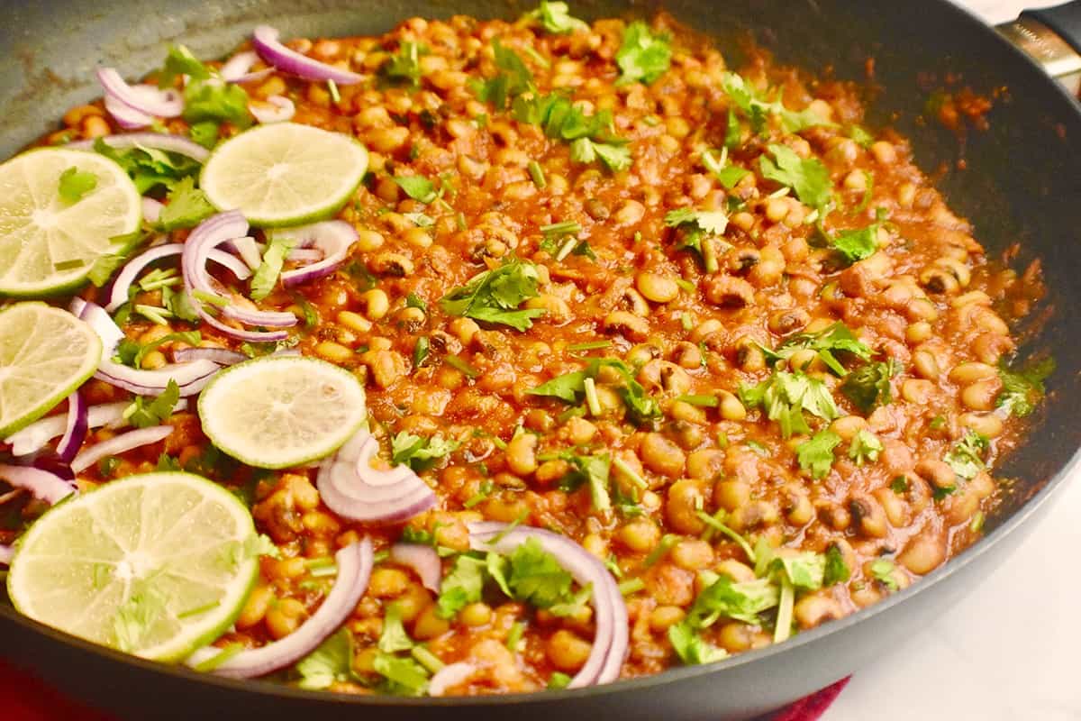 Black pan with cooked black eyed peas (Punjabi lobia) recipe, garnished with red onions, sliced lemons and cilantro.