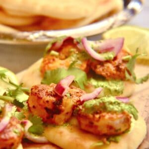 A white countertop with plates full of tandoori shrimp cooked on a grill pan and served with mini naan, pickled onions, cilantro chutney and wedges of limes.