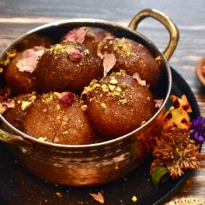 gulab jamun garnished with pistachios and rose petals in a copper serving bowl on a black plate with flowers.