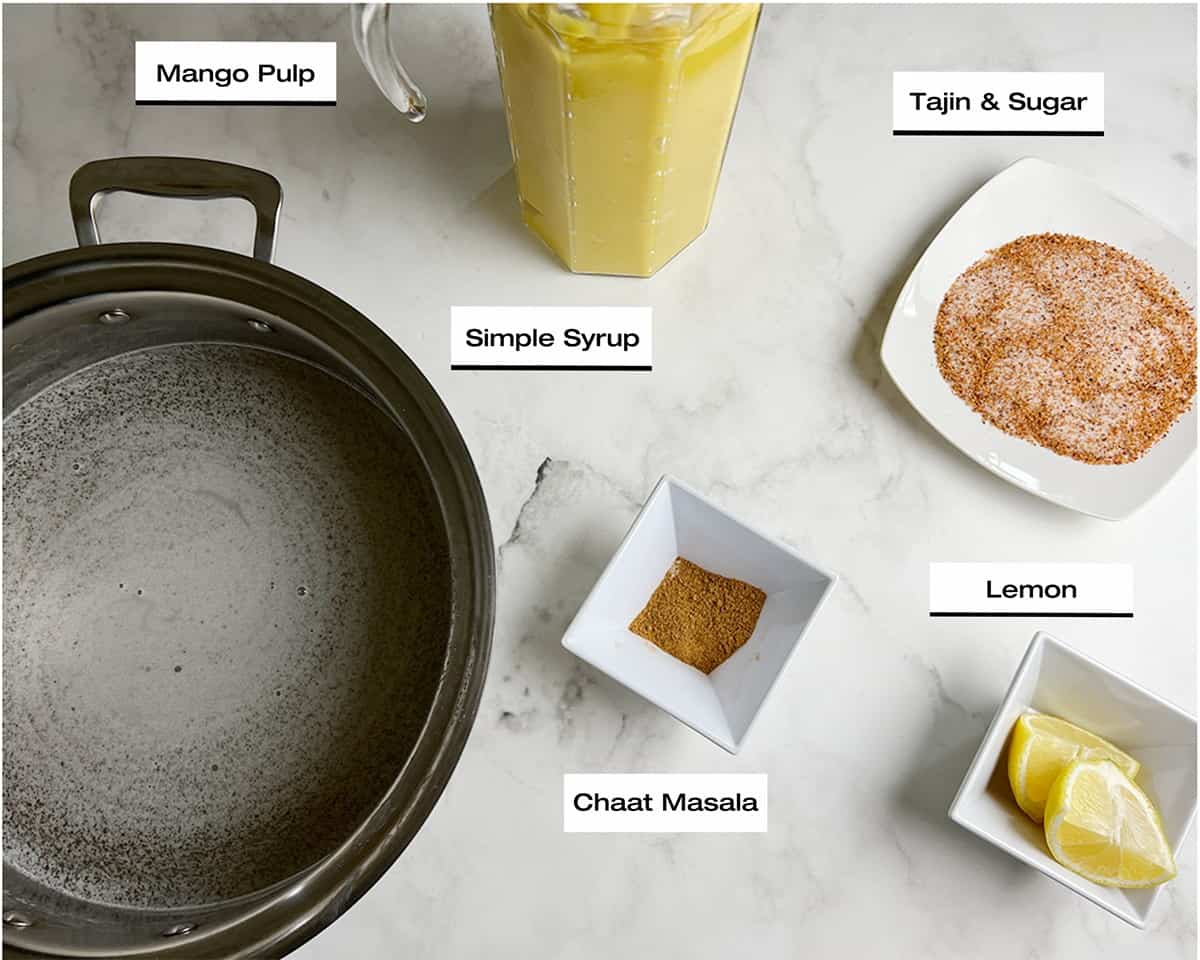 The ingredients for a mango juice recipe; prepared mango puree, a simple syrup, chaat masala, lemons and tajin and sugar, rest on a white marbled kitchen counter.