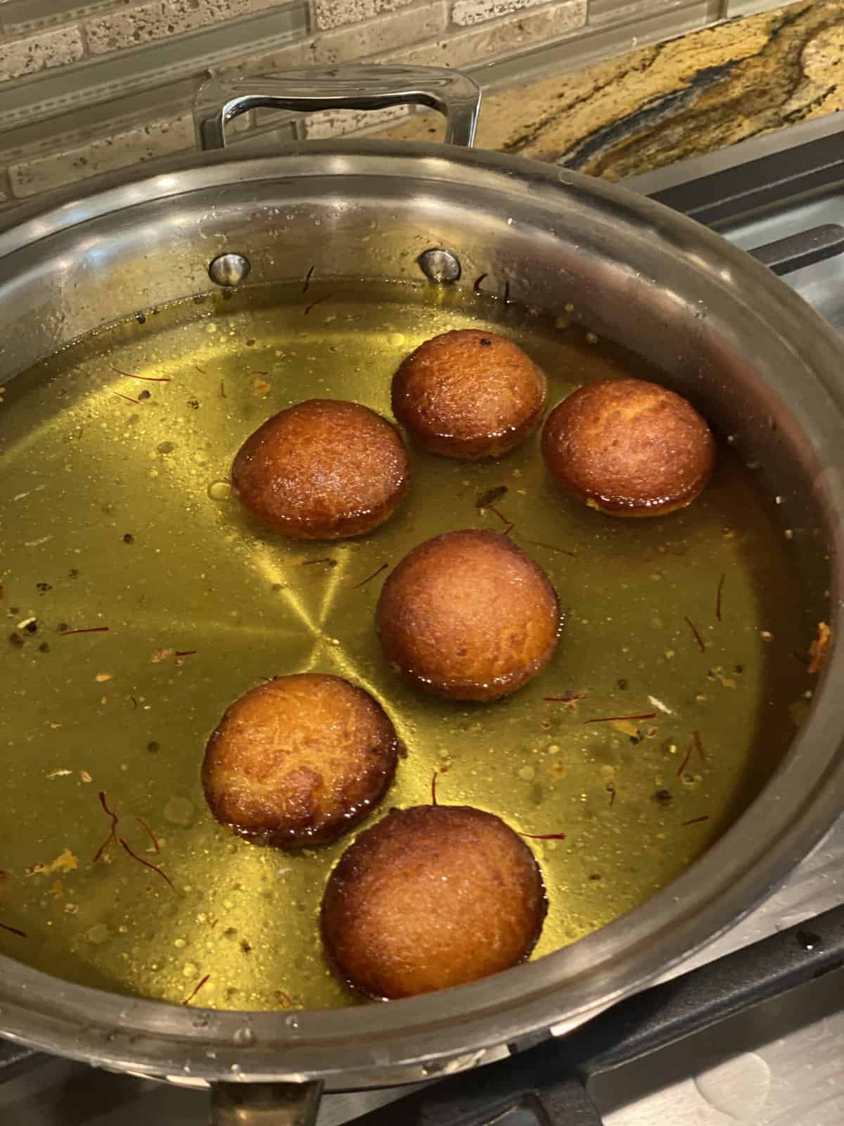 A silver pan filled with sugar syrup and fried gulab jamun.