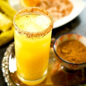A glass of mango juice rimmed with tajin and sugar rests on a silver tray alongside a small bowl of chaat masala.