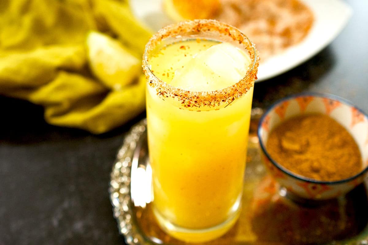A glass of mango juice in a tall glass full of ice and rimmed with tajin and sugar rests on a silver tray, besides a bowl of chaat masala, with lemons in the background.
