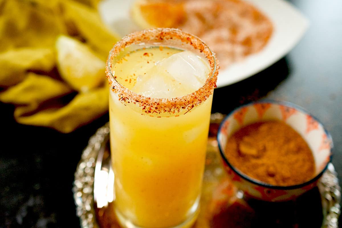 A glass of mango juice rimmed with tajin and sugar rests on a silver tray alongside a small bowl of chaat masala.