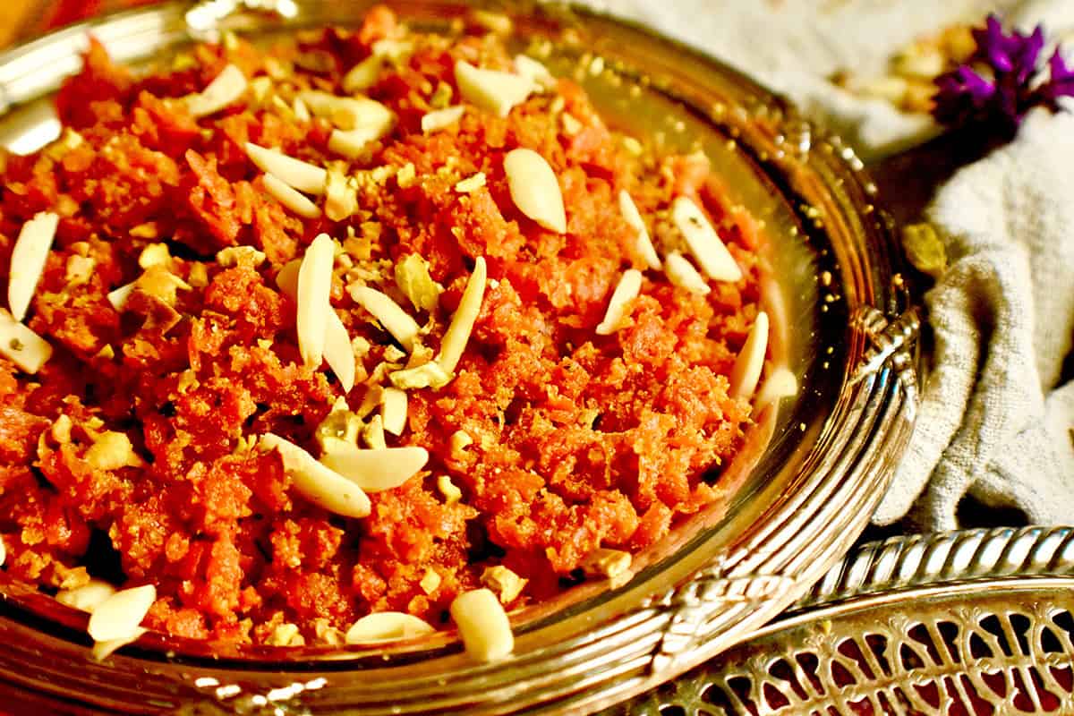 A silver dish filled with carrot halwa, garnished with almonds and pistachios, is resting on a grey napkin with a purple flower in the background.