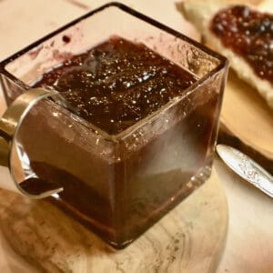 A glass jar filled with plum jam on a wooden board with a spoon in the jar.