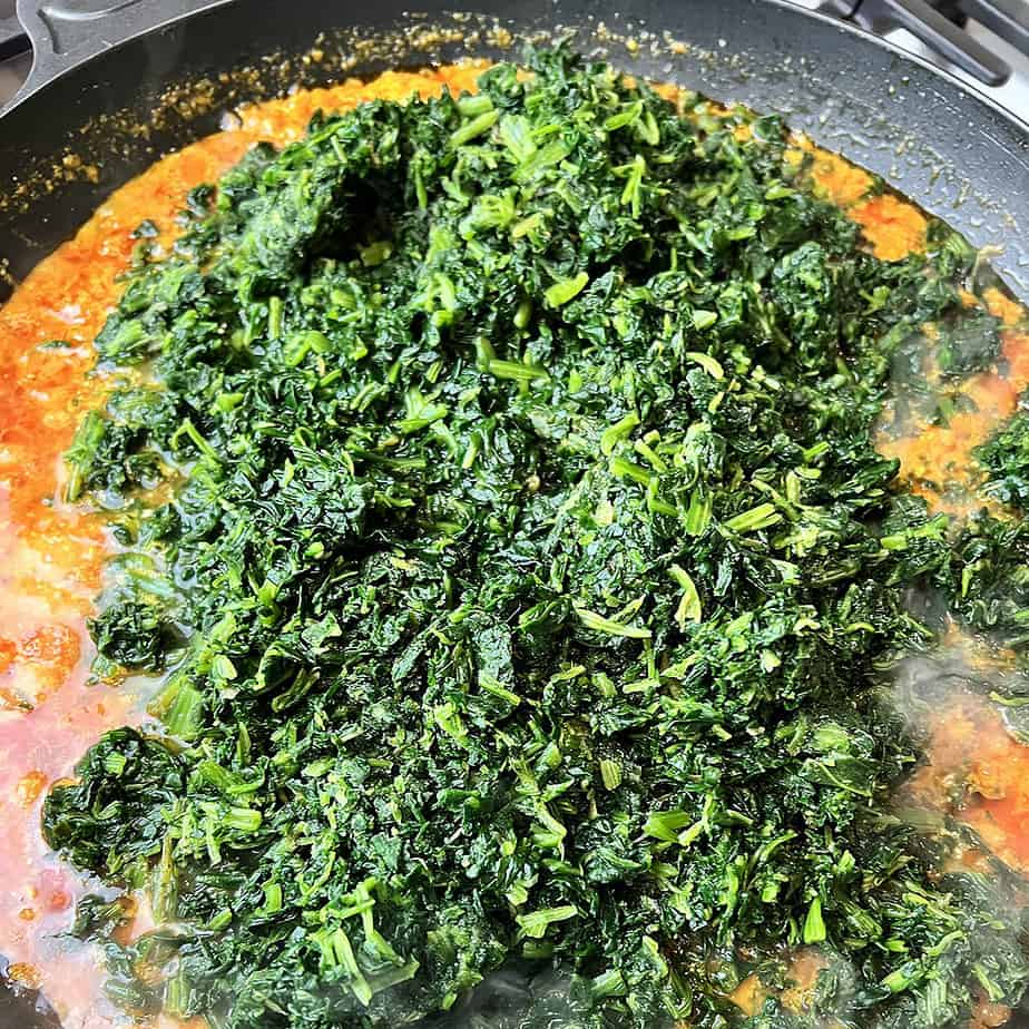 frozen palak being added to a curry base in a black saute pan.