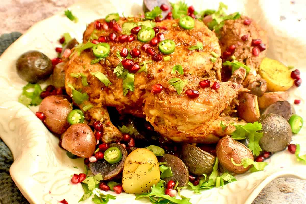 Roast chicken is plated in a white platter, garnished with pomegranate seeds and served with roasted multi colored potatoes. 