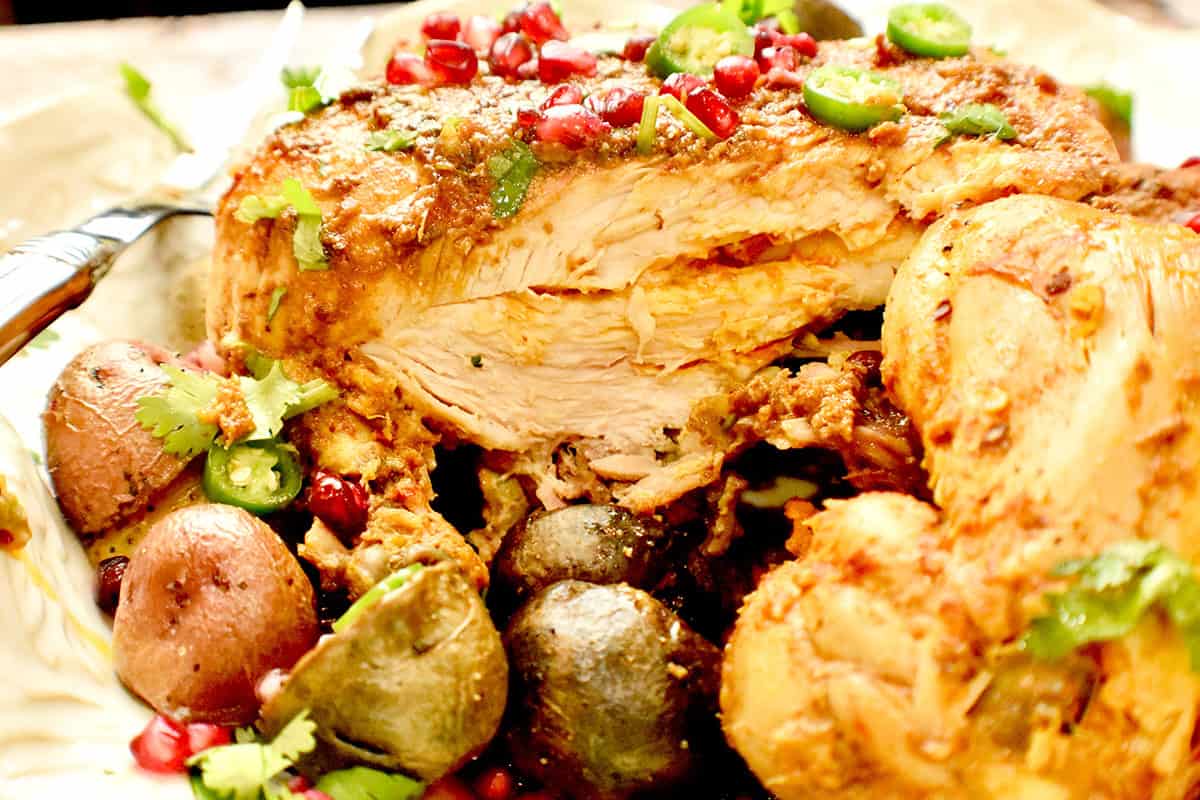 A partially cut Pakistani steamed chicken roast in a white platter with roasted potatoes and a garnish of pomegranate seeds.