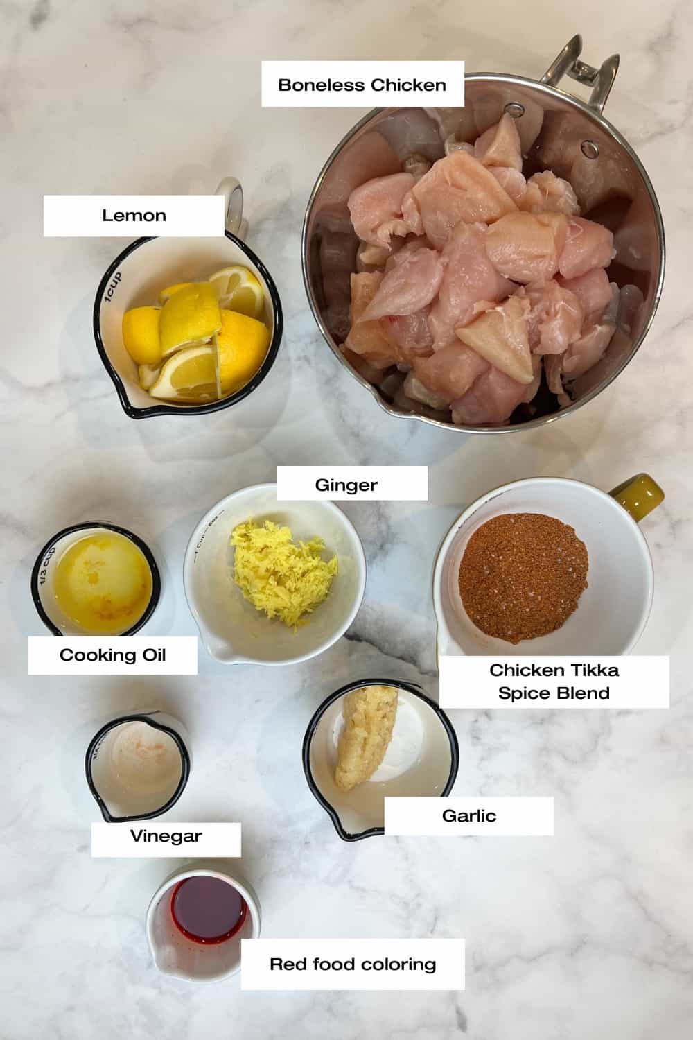 white kitchen counter with bowls filled with boneless chicken, lemons, cooking oil, white vinegar, garlic, ginger, chicken tikka spice blend and red food color.
