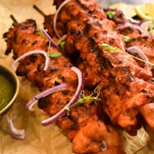 Brown parchment with cooked chicken tikka boti on skewers with green chutney, onions and micro greens.