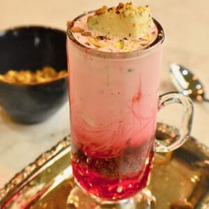 An off white countertop with a blue bowl full of pistachios and a tall glass filled with Falooda (Pakistani ice cream dessert)