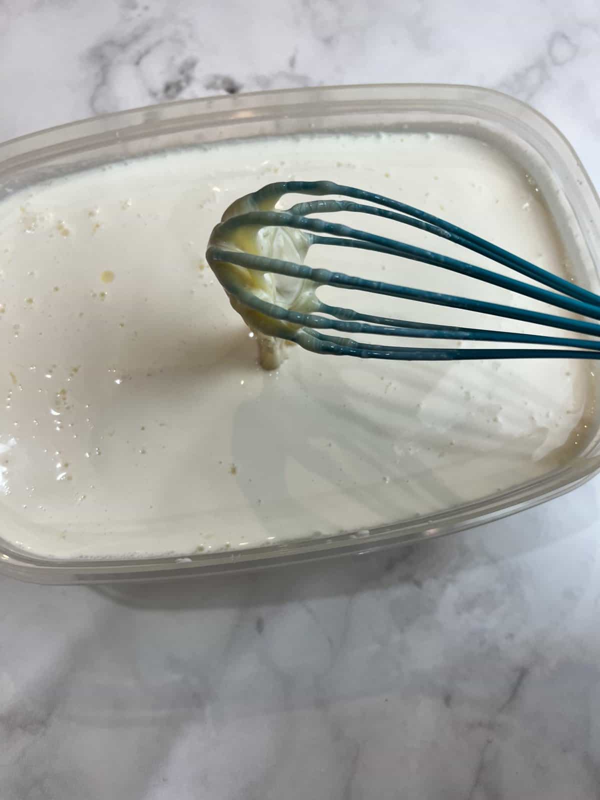 A white countertop with a tupperware container filled with all of the liquid ingredients for kulfi being mixed together with a blue whisk.
