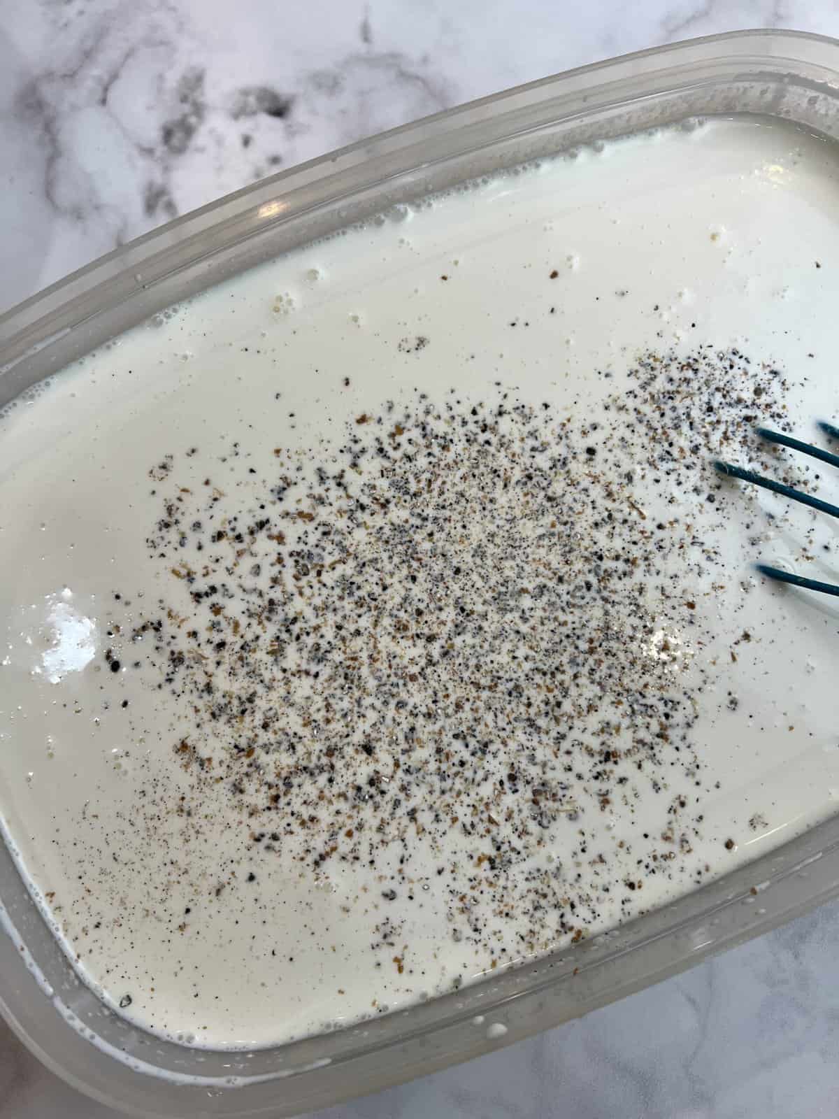 A white countertop with a container filled with a kulfi mixture and crushed cardamom being stirred in.