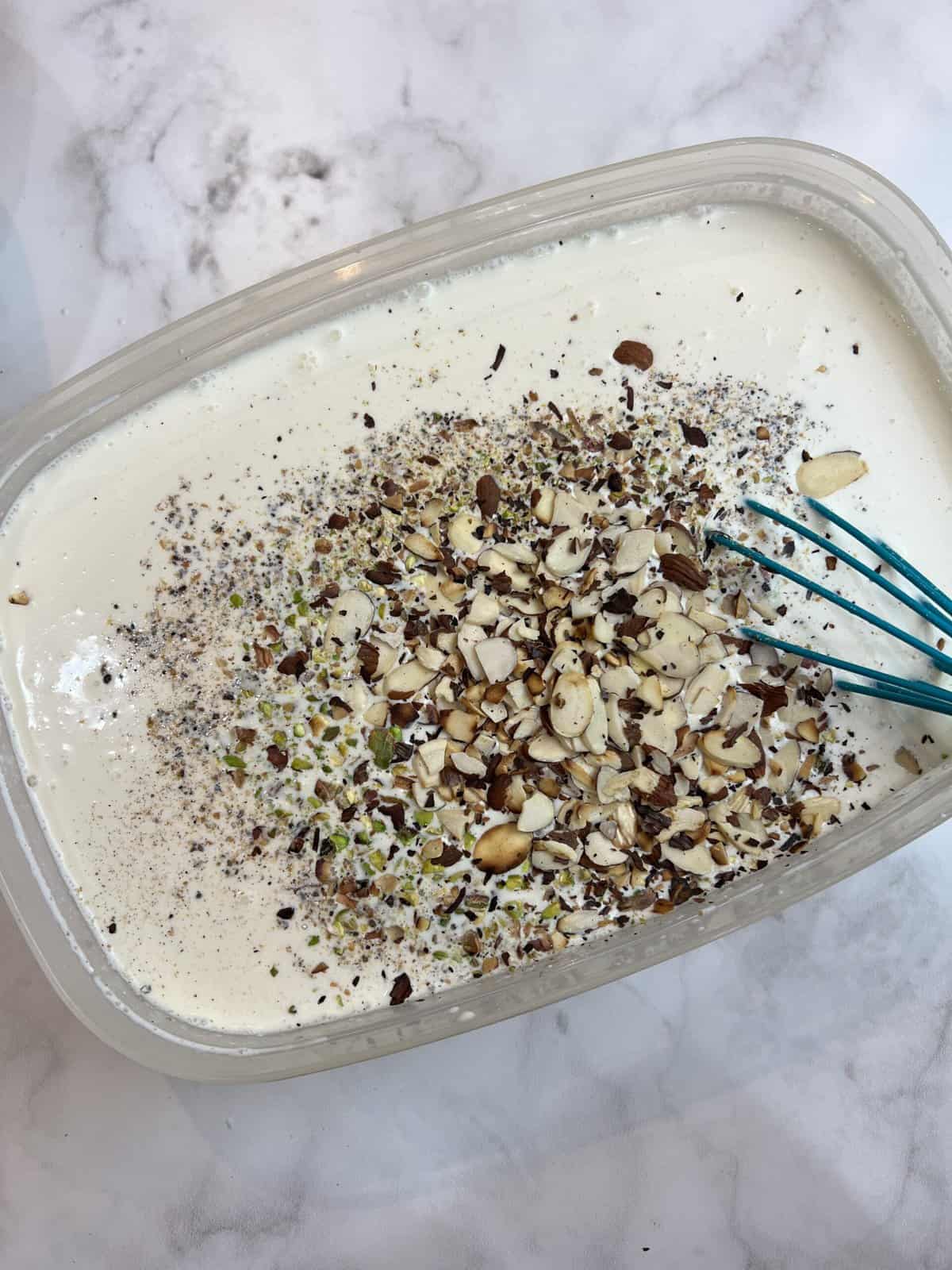 A white countertop with a container filled with a kulfi mixture and roasted and chopped nuts being stirred in.