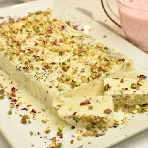 A white platter with kulfi (pakistani ice cream) topped with pistachios and rose petals, with a glass jug filled with rose milk and rice noodles.