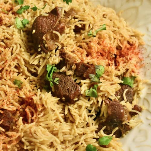 Mutton Pulao (rice pilaf with goat meat) garnished with cilantro and sprinkled with red chili powder is plated in a white ceramic dish.