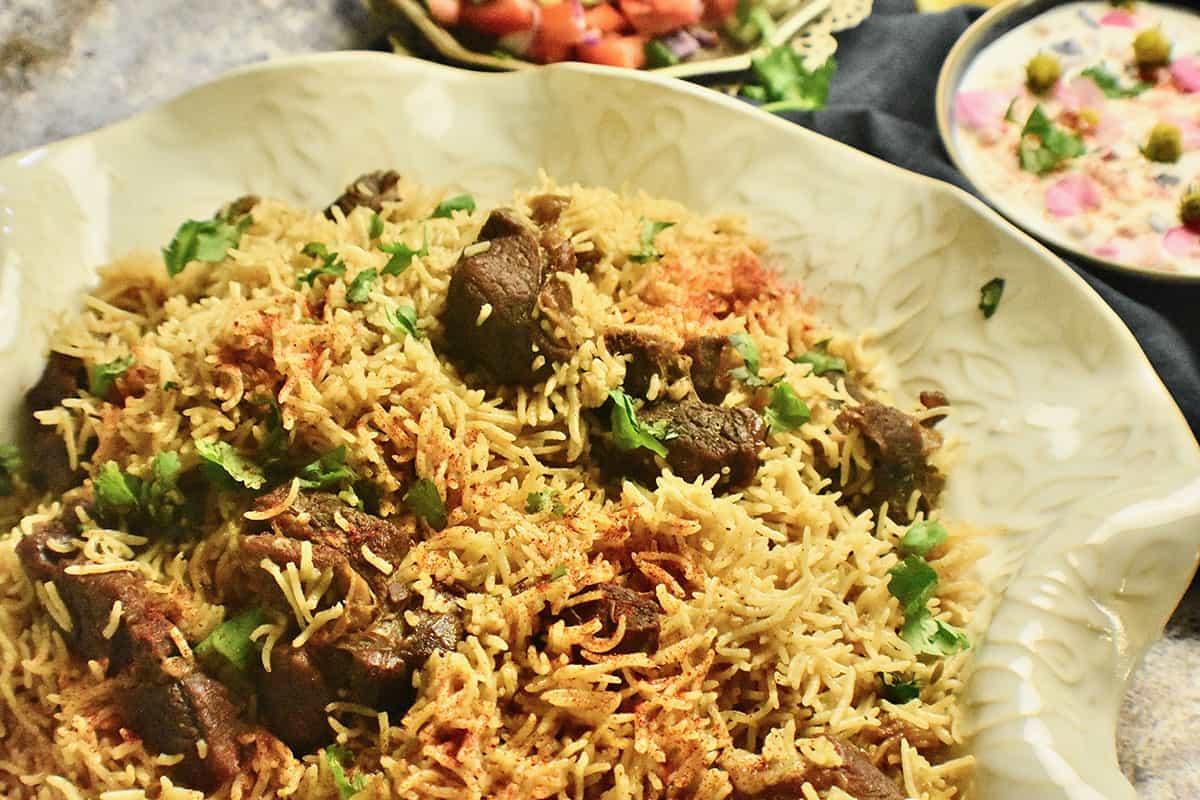 Mutton Pulao (rice pilaf with goat meat) garnished with cilantro and sprinkled with red chili powder is plated in a white ceramic dish and served with a chopped salad and yogurt dip (raita).