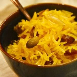 A blue bowl with a spoon resting on a white napkin is filled with Zarda Recipe (Pakistani sweet rice) with nuts, raisins, cardamom and cloves.
