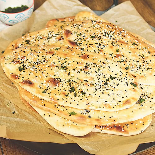 Flatbread made using an easy naan recipe is resting on brown parchment paper straight out of the oven with bowls filled with toppings for the naan.