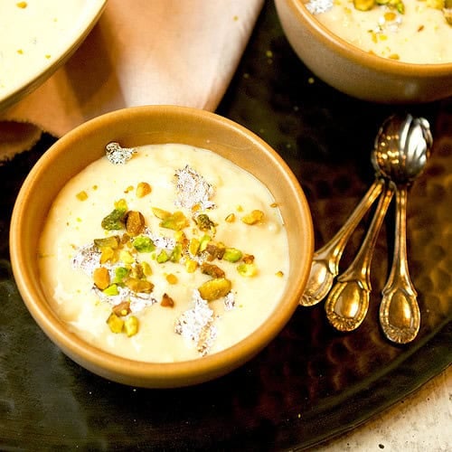 A small earthen bowl is filled with rice kheer that is garnished with pistachios and silver leaf.