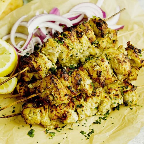 Chicken malai tikka skewers accompanied with red onions and lemon wedges are resting in a platter lined with brown paper.