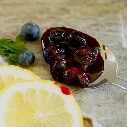 A spoon full of a freshly cooked blueberry compote is resting on a kitchen counter, surrounded by lemons, fresh blueberries and some mint.