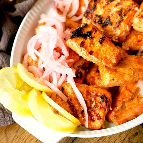 A white platter is filled with cooked tandoori fish, pickled onions and sliced lemons.