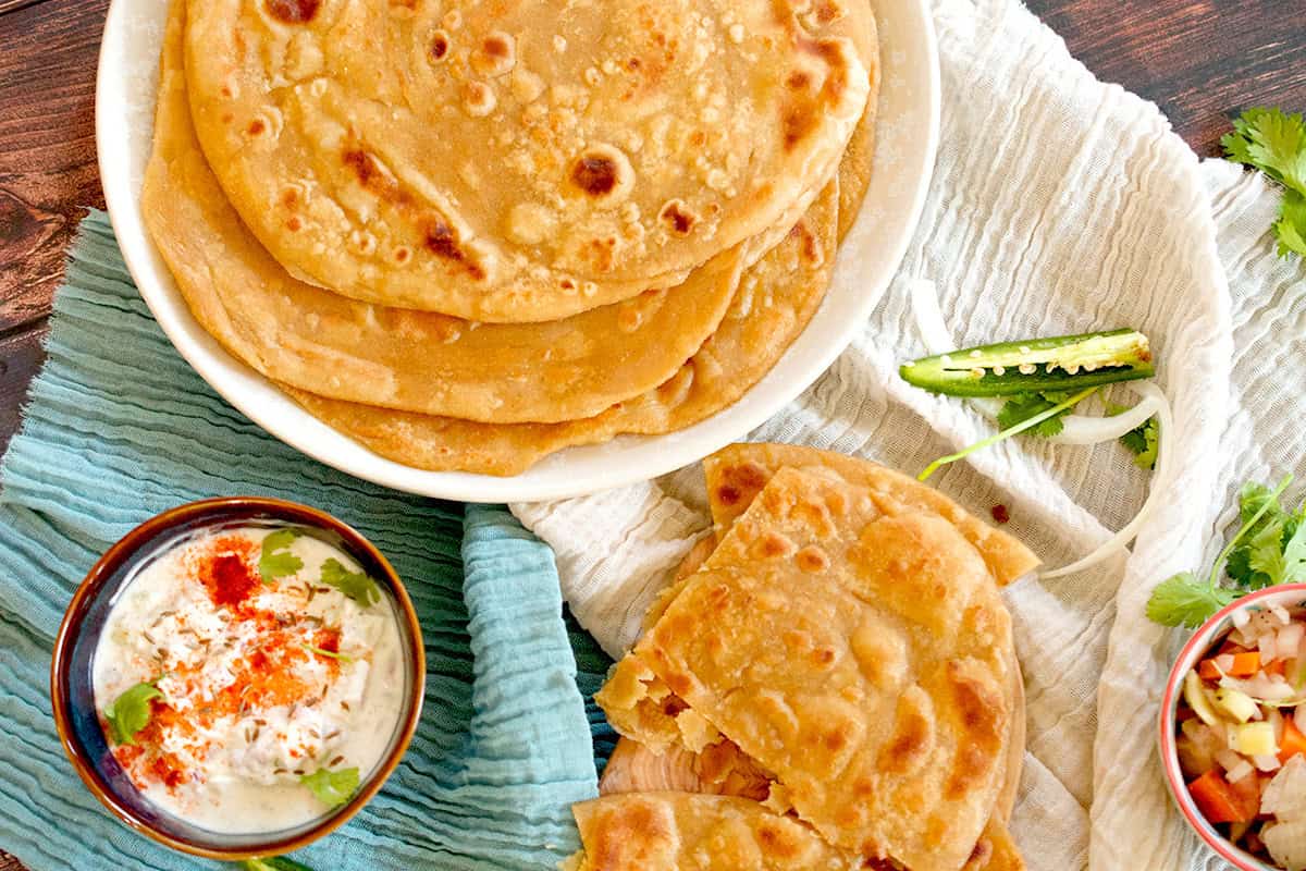 Cooked parathas are plated and served with some raita and green chilies.