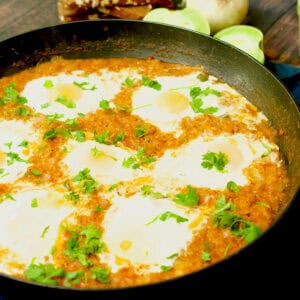 Poached eggs in a green tomato sauce have been cooked in a large black frying pan and are being served with some bread on a wooden table.