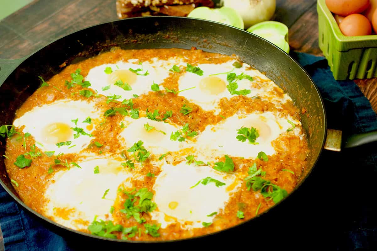 A cooked green shakshuka recipe in a black frying pan is resting on a wooden table with green tomatoes, bread and eggs in the background.