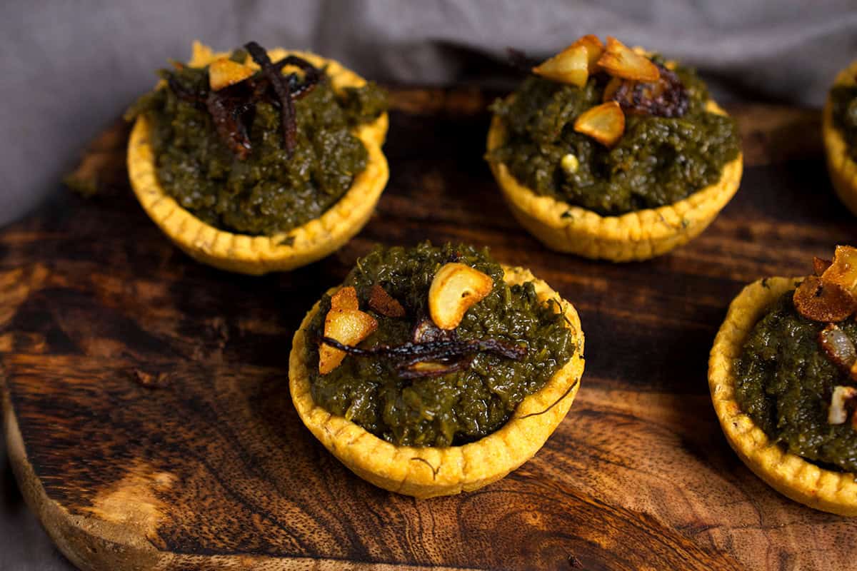 makki ki roti tarts filled with mustard greens are served on a dark wooden board.