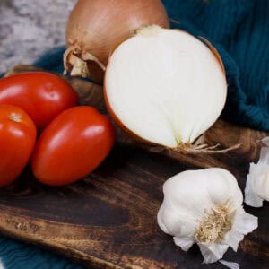 Onions, tomatoes and garlic are lying on a wooden board in prep to make a curry.