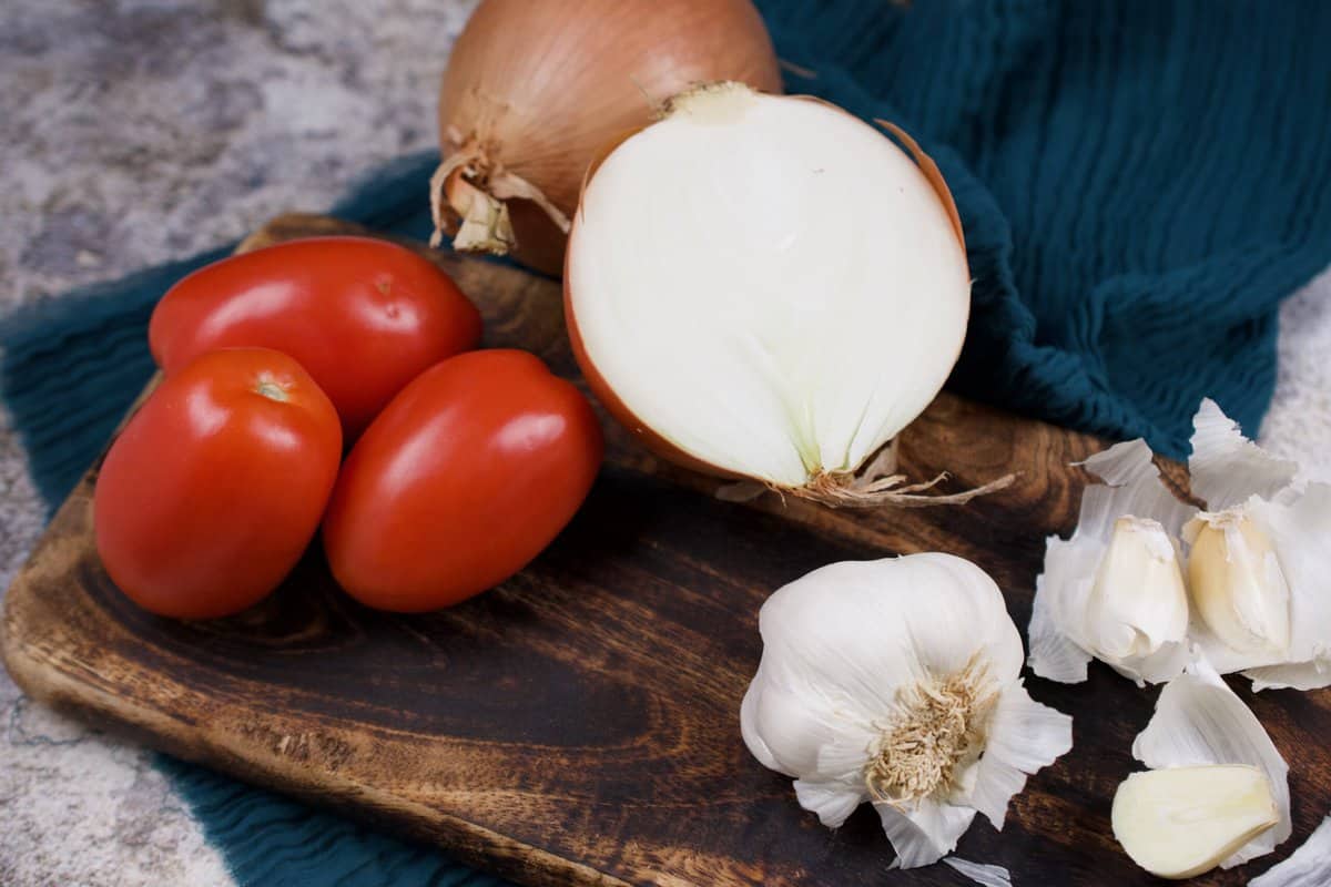 Onions, tomatoes, and garlic are resting on a wooden board to be prepped for a curry.